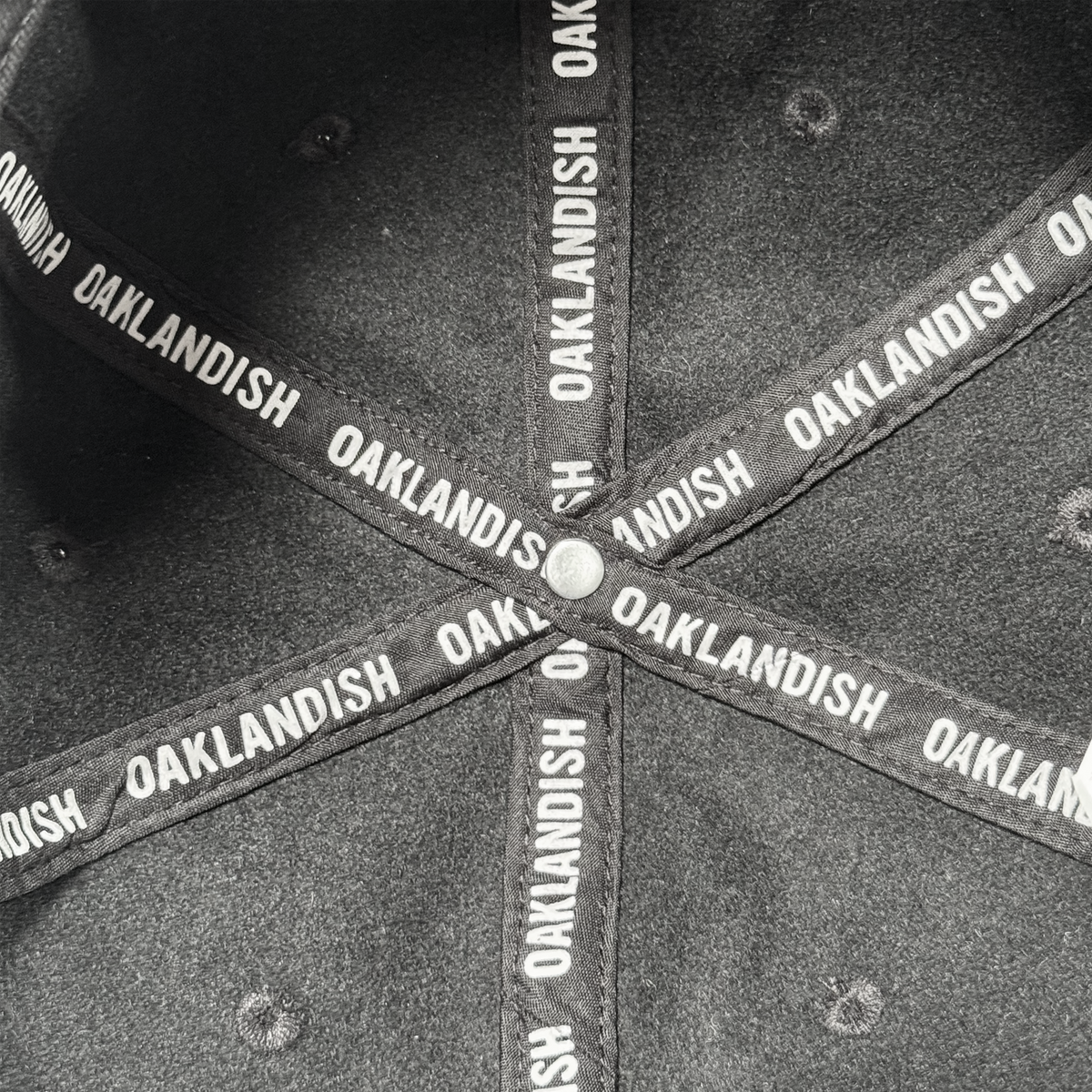 Close-up of the inside of an Oakland Ballers B Strapback hat, showing crossed black fabric strips with “OAKLANDISH” in white uppercase letters and a metal button at the intersection. Brand: Oakland Ballers.
